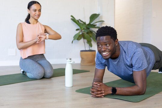 African American pair in gymwear holding plank checking trackers on green mats at home with bottle