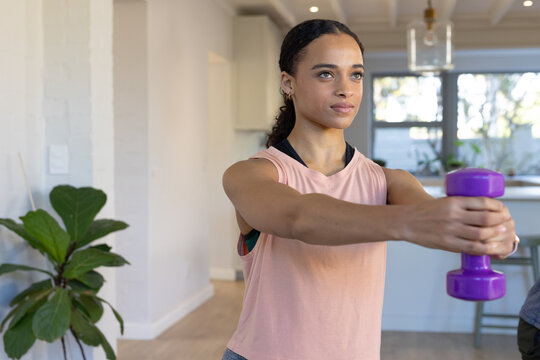 African American woman in sleeveless pink top holding purple dumbbell and exercising in kitchen