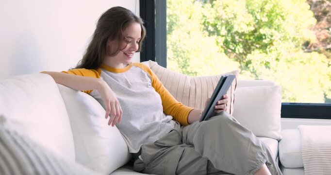 Adult woman wearing white tee yellow sleeves reacting to tablet, swiping, smiling on sofa by window