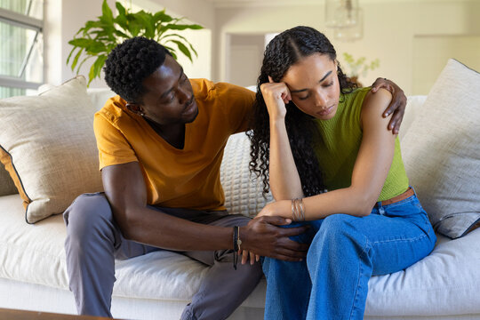 African American couple sitting close on light sofa in living room, man leaning, bracelets visible