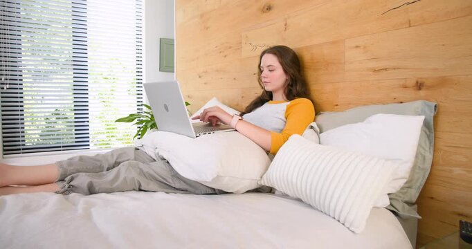 Female working on silver laptop on bed reacting to screen, laughing, hiding behind striped pillow