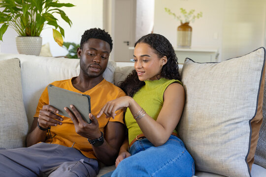 African American couple sitting on sofa in casual clothing holding tablet and pointing at screen