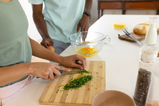 African American couple chopping herbs and cracking eggs at kitchen island with knife, mixing bowl