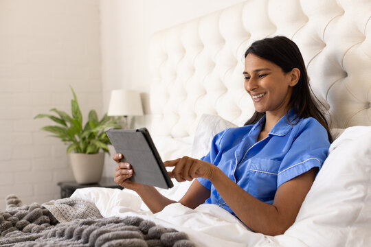 Indian woman in blue pajamas sitting up in bed holding tablet and tapping screen, copy space