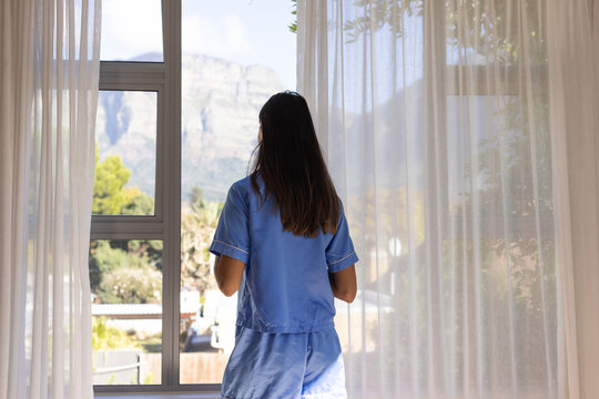 Female standing at home in blue pajamas, facing paned window with sheer curtains and mountain view