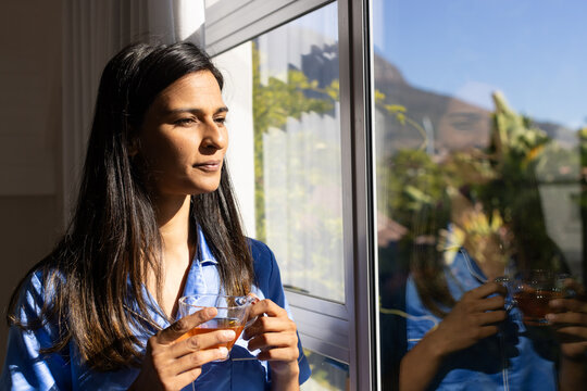 Indian woman in blue satin pajamas standing at large window holding clear cup gazing, copy space