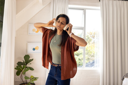 Indian woman standing in loft room wearing rust shirt and olive top, holding headphones, listening
