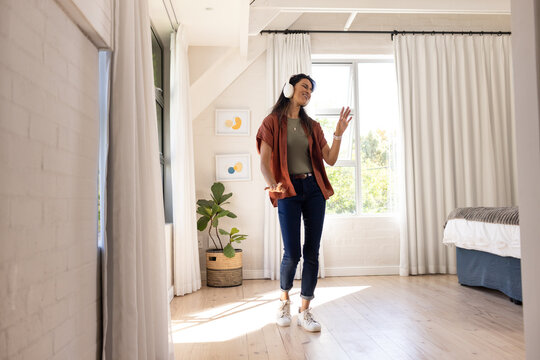 Indian woman standing and swaying in bright bedroom near window with white headphones