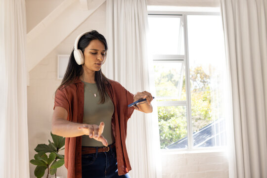 Indian woman wearing over-ear headphones holding smartphone, gesturing while listening at home loft