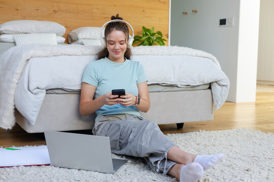 Adult female sitting on shag rug in bedroom, wearing headphones and using smartphone and laptop