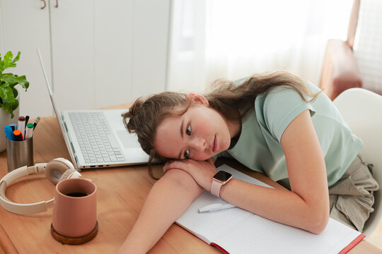 Woman resting head on folded forearms across notebook at wooden desk with laptop, mug, smartwatch
