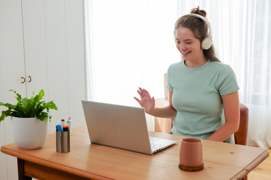 Adult woman wearing green top, beige headset, waving at laptop in home-office with mug plant pens