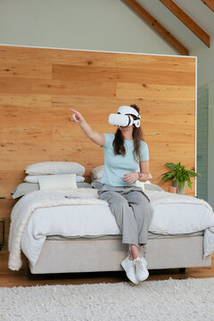 Woman sitting on edge of bed in bedroom wearing white VR headset reaching toward phone