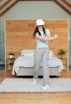 Woman standing centered in bedroom wearing white VR headset and interacting with virtual objects