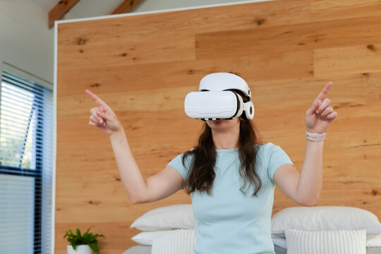 Adult woman standing in bedroom wearing white VR headset, pointing and interacting with smartwatch
