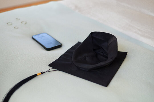 Black graduation cap is resting on pale bed near smartphone and metallic rings, soft light