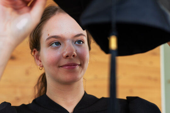 Woman holding graduation cap with tassel near head, wearing gown and hoop earrings in hall