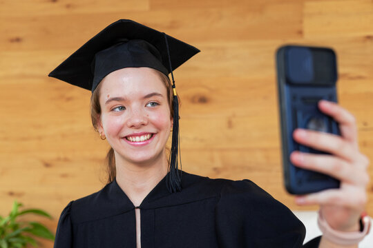 Female graduate aged 18-25 holding smartphone, taking selfie by wood wall with mortarboard and gown