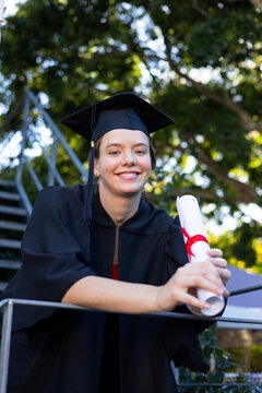 Female graduate standing on campus metal rail holding red-ribbon diploma wearing cap and black gown