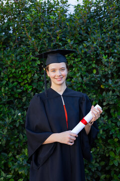 Adult female in black gown and cap standing before green hedge holding diploma with red ribbon