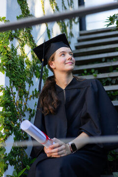 Female graduate sitting on exterior stairway holding rolled diploma, wearing gown and mortarboard