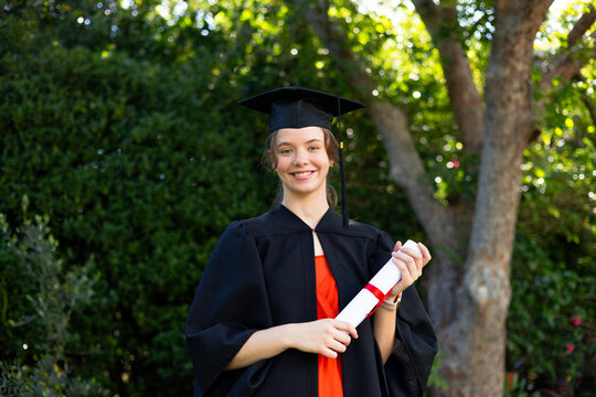 Woman in her 20s standing in sunlit campus garden wearing gown and mortarboard holding diploma