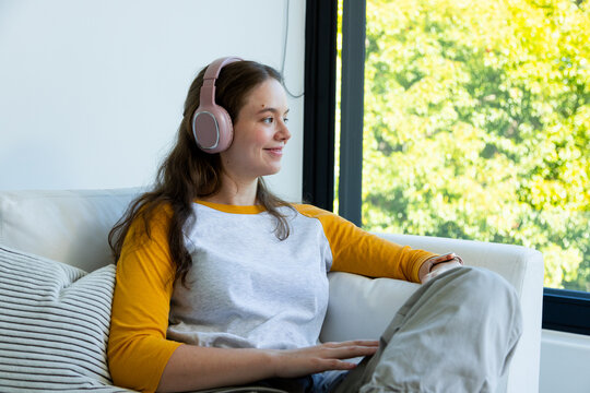 Woman sitting on white sofa at home wearing pink headset mustard raglan shirt looking out window