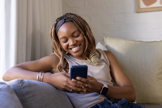 Adult woman reclining on light couch by window holding blue smartphone wearing headband and rings