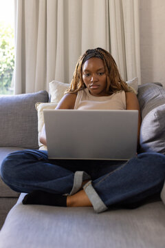 African woman sitting cross-legged on grey sofa using silver laptop with patterned headband