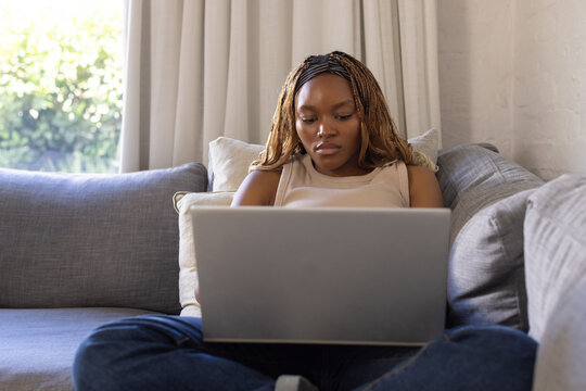 African woman sitting on grey sectional sofa using silver laptop at home wearing sleeveless top
