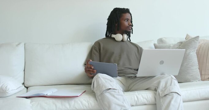 Adult African American man glancing right, alternating tablet with laptop while working on sofa