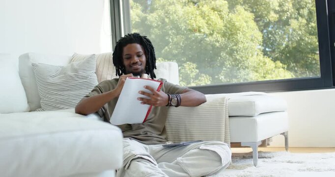 African American man sitting on rug in living room paging white notebook red spine studying smiling