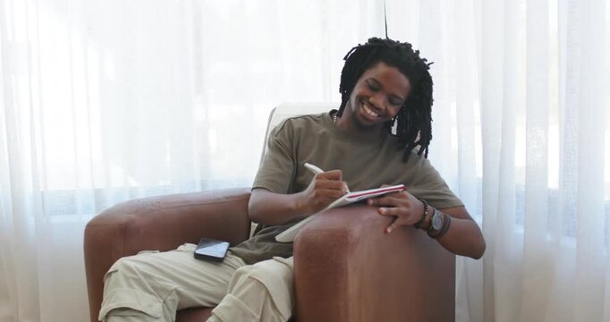 African American man starting sketching on pad with pen in armchair by bright window, phone resting