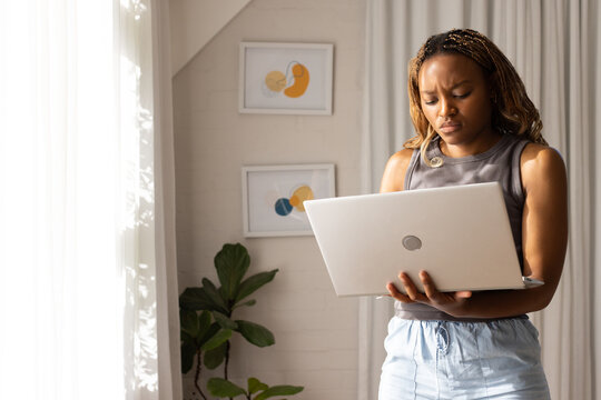 African woman standing near window at home holding silver laptop in gray tank shorts, copy space