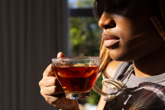 African American woman wearing grey tank holding glass cup of tea sipping near window, copy space