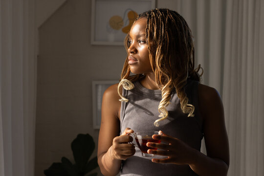African American woman standing near sunlit window holding glass cup in sleeveless top, framed art