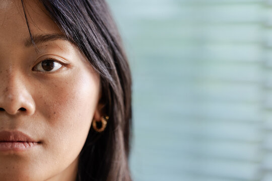 Asian woman gazing at window blinds in room, showing gold hoop earring, copy space