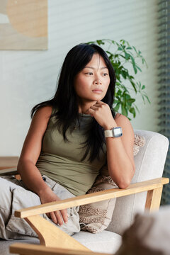 Asian woman in thirties sitting in wooden armchair, resting elbow, gazing right, wearing smartwatch