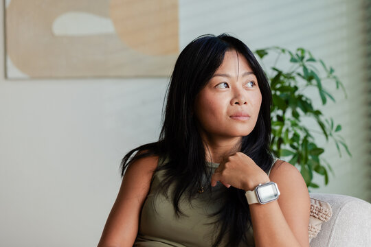 Asian woman sitting on light sofa at home, wearing olive-green top and white smartwatch