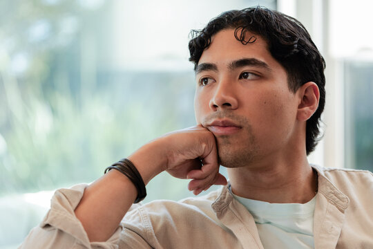 Asian man sitting on sofa near window, wearing beige shirt, wrap-bracelet, gazing right, copy space