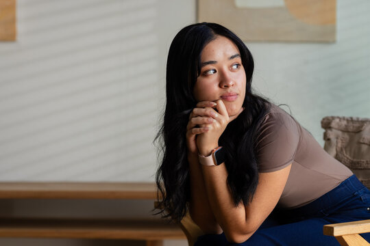 Woman sitting on wooden chair in living room, leaning forward, wearing taupe top and smartwatch