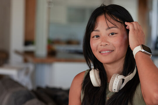 Asian woman sitting on right of living room, tucking hair, wearing headphones, smartwatch, pendant