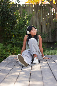 Asian woman sitting on wooden deck in backyard garden wearing white headphones and olive-green top