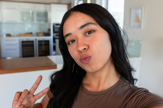 African American woman taking selfie in kitchen with ovens, in brown top, holding peace sign