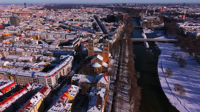 Aerial winter view of St. Maximilian Church and Reichenbach Bridge over the Isar River in Munich, Germany. Scenic drone panorama of snowy Bavaria and historic city center.