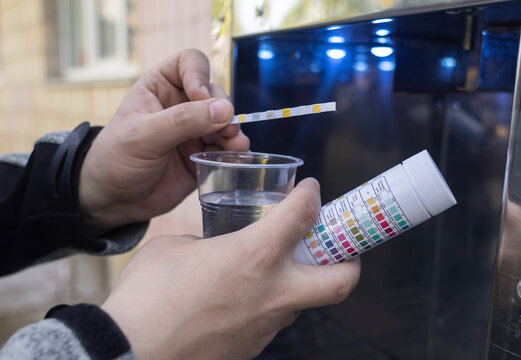 Man checking water quality at a public vending machine using a test strip. Analysis of purified drinking water in a plastic cup. Safety, pH, and hardness control of urban water supply.