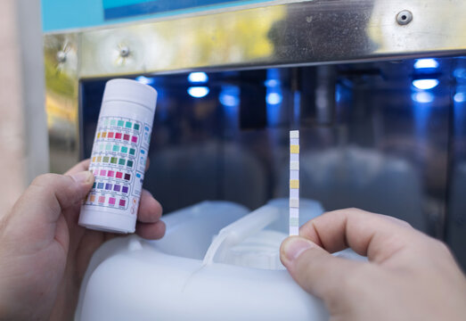Man checking water quality at a public vending machine using a test strip. Analysis of purified drinking water in a plastic cup. Safety, pH, and hardness control of urban water supply.