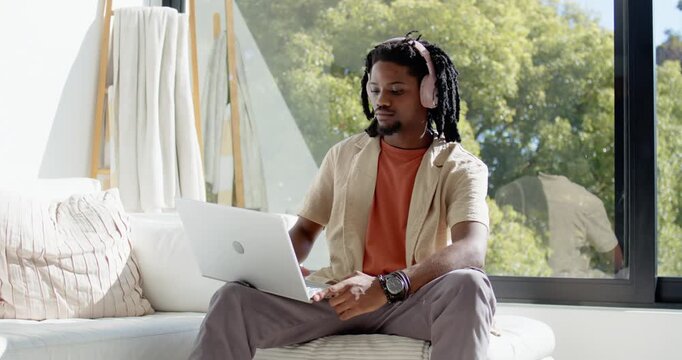 African American man wearing headphones working on silver laptop, typing, leaning on sofa by window