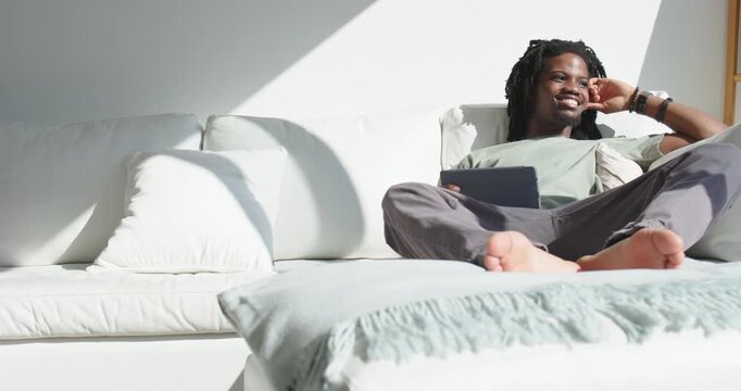 Man sitting on right sofa, supporting head, nudging tablet, slowly smiling at screen, copy space