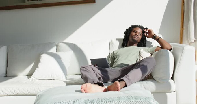 Adult man reclining on white sofa at home, resting tablet, adjusting hair, turning toward sunlight